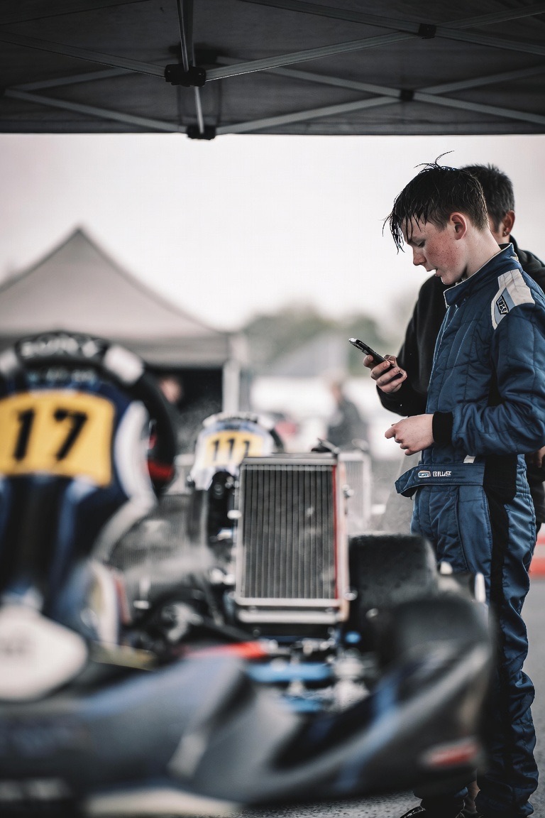 Young kart driver under the team awning between sessions, checking the next call on a phone beside the kart